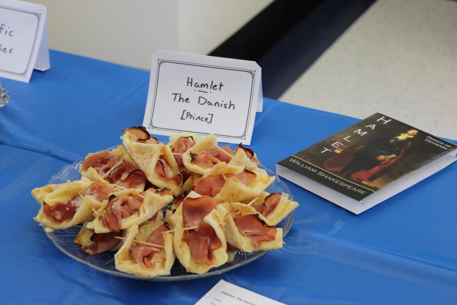 A plate is piled up with some meat danishes. A paper placard is placed near the dish with the words Hamlet The Danish [Prince] written on it. Also nearby is the actual Hamlet book by William Shakespeare. The book, the dish and the placard are all placed on the same table that has a bright blue sheet covering it.