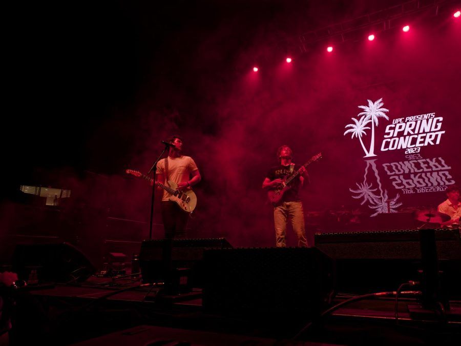 Two guitarist perform on stage flooded with red lights and copious amounts of smoke, giving the picture an overall red tint. The picture is taken from a lower angle from the right, facing the stage. One of the performers is directly in front of the mike, wearing a white T shirt with black pants and holding a white guitar. His head is turned towards the other musician a little behind him who is wearing a black shirt and light coloured pants and holding brownish looking guitar. The stage backdrop consists of the message UPS PRESENTS SPRING CONCERT 2023 alongside an illustration of two tropical trees situated on the messages left.